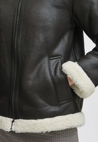 Jachetă din piele neagră cu închidere cu fermoar, având margine din blană crem la manșete și la tiv, precum și buzunare laterale. Exterior texturat.