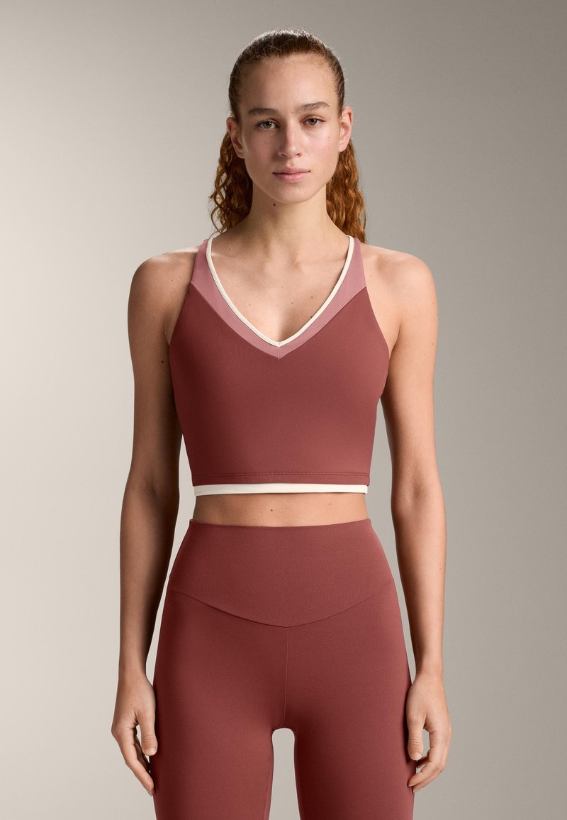 Donna con capelli ricci raccolti in una coda alta che indossa un reggiseno sportivo bicolore malva e leggings a vita alta abbinati, su uno sfondo neutro.