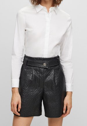 Valkoinen pitkähihainen nappilista, joka on sullottu korkeavyötäröisten mustien, tekstuuripintaisesti nahkaisten shortsien sisään, jossa on vyö ja hopeinen solki.