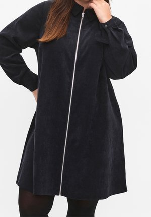 Vrouw die een loszittende zwarte corduroy jurk draagt met lange mouwen en een zilveren ritssluiting aan de voorkant, poseert met één hand op haar heup.