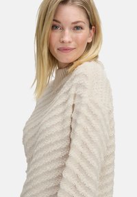 Blonde Frau mit glattem Haar, die einen strukturierten cremfarbenen Strickpullover trägt und mit einem leichten Lächeln in die Kamera schaut, vor einem weißen Hintergrund.