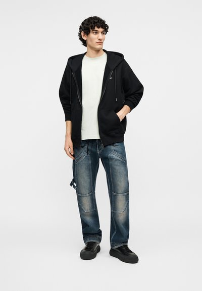 Άνδρας που φοράει μαύρο φούτερ με φερμουάρ, λευκό αντικείμενο μπλουζάκι, μπλε patchwork τζιν και μαύρα αθλητικά παπούτσια, στέκεται με το ένα χέρι στην τσέπη.