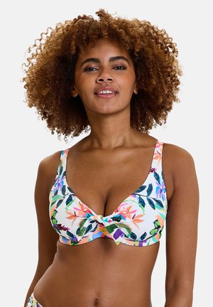 Vrouw met krullend haar die een witte bikini top met bloemmotief draagt, glimlacht en recht vooruit kijkt tegen een effen achtergrond.
