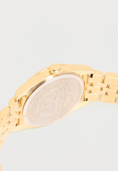 Ρολόι χειρός σε χρυσό τόνο που δείχνει την πίσω πλευρά χαραγμένη με κεφάλι Μέδουσας και αλυσίδα από μέταλλο.