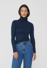 Pull à col roulé côtelé navy avec des manches longues, associé à un jean bleu taille haute, présentant un design classique à cinq poches et une fermeture à bouton.