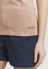 Ljusrosa bomulls t-shirt med rundad nederkant, prydd med en liten blå "OXMO" logotyp och en trähjärta-detalj. Matchad med marinblå shorts.