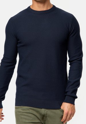 Man wearing a dark blue textured long-sleeve sweater with ribbed cuffs, paired with olive-green pants against a plain background.