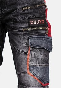Donkergrijze denim cargobroek met rode accenten. Beschikt over meerdere met rits afgesloten zakken en een gestructureerde afwerking. Inclusief een patch met tekst.
