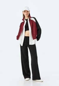 Röd och svart zip-up jacka med vita detaljer, bärs över en svart cropped topp, ihop med lösa svarta byxor och vita skor, mot en enkel bakgrund.