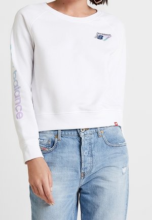 Biała krótka bluza z logo New Balance na piersi i rękawie, noszona z jasnoniebieskimi, biodrówkami dżinsami.