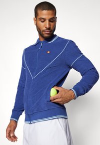 Blå zip-up treningsjakke med hvite kontrastlinjer og ribbestrikkede mansjetter, med en liten logo på brystet og en lomme som holder en tennisball.