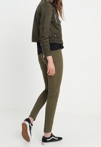 Giacca in suede verde oliva corta con zip nera, abbinata a pantaloni slim-fit verde oliva e sneaker nere Vans.