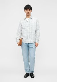 Άνδρας με ανοιχτόχρωμο πουκάμισο με καρό, ανοιχτό μπλε τζιν, μαύρα παπούτσια, κρατώντας μικρή ταμπά τσάντα στο δεξί χέρι, στέκεται μπροστά σε λευκό φόντο.