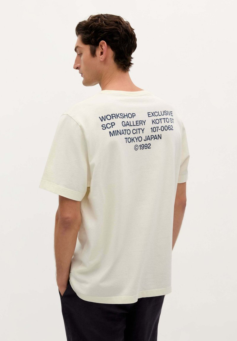 Tricou scurt cu mâneci, de culoare crem, fabricat din bumbac, cu croială lejeră. Partea din spate prezintă un text bleumarin în diverse fonturi, incluzând "WORKSHOP EXCLUSIVE" și "©1992."