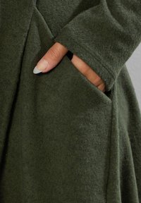 Olivgrön tyg med en mjuk, filtliknande textur. Har en sidficka och en avkopplad passform med långa ärmar och en rundad nederkant.