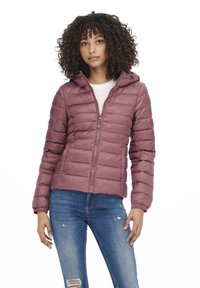 Veste à capuche, en mauve, avec un rembourrage, comportant une fermeture éclair à l'avant, des poignets élastiques et une silhouette ajustée. Portée par-dessus un haut léger et un jean déchiré.