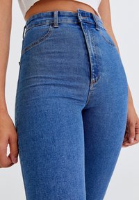 Jean en denim bleu taille haute avec une texture lisse, présentant un léger effet délavé, des coutures contrastantes et des détails de poche avant.