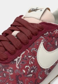 Crvene tenisice s uzorkom i teksturiranim bordo vezicama te bijelim detaljima. Imaju istaknutu Nike oznaku sa strane i podstavljeni ovratnik.