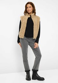 Gilet in finta pelliccia beige, camicia nera a maniche lunghe, pantaloni grigi e stivali neri alla caviglia. Tessuto morbido, design stratificato, stile casual.