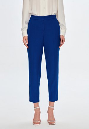 Kobieta ubrana w dopasowane, królewskie niebieskie spodnie sięgające kostek z wysokim stanem, połączone z białymi sandałami na obcasie i kremową bluzką.