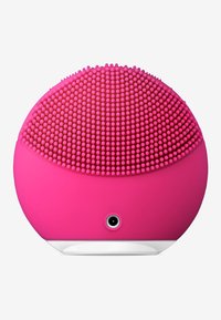 Brosse faciale en silicone rose, de forme ronde, avec des nodules texturés et surélevés d'un côté et une base lisse avec une petite prise audio.