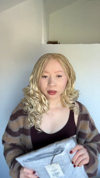Geflochtenes blondes Haar, trägt eine gestreifte braune und beige Strickjacke, hält ein graues Paket mit einem Anhänger. Hintergrund ist eine einfarbige helle Farbe.