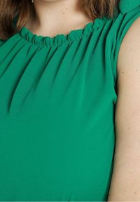 Top verde senza maniche con scollatura arricciata e tessuto leggero e morbido. Superficie texture con pieghe sottili vicino alle spalle.