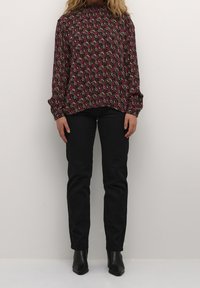 Langärmlige Bluse aus dunklem Stoff mit einem rosa und goldenen Blumenmuster; lockere Passform, hoher Kragen und geraffte Manschetten, kombiniert mit schwarzen Hosen.