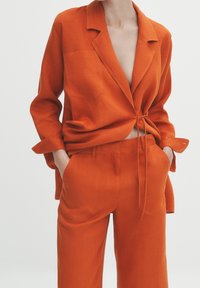 Oranje linnen blazer met een strik in de taille, gecombineerd met bijpassende wijde broekspijpen. Heeft een enkele borstzak en opgerolde mouwen.