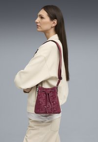 Bolso de cuero burdeos tipo bucket con patrón geométrico grabado, correa de hombro ajustable y cierre de cordón. Textura suave, diseño compacto.