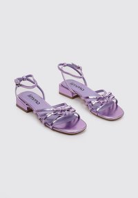 Sandalias de tacón metálicas moradas con punta cuadrada, correa ajustable en el tobillo y detalles decorativos de lazo en la parte delantera. Tacón en bloque pequeño.