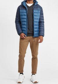 Blå puffjakke med hætte, med quiltet design, båret over en grå sweater. Kombineret med brune bukser og hvide sneakers.