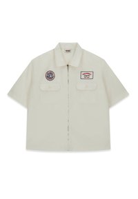 Camicia a maniche corte di colore crema, realizzata in cotone, con due tasche sul petto e toppe ricamate, chiusura con zip e colletto piatto.