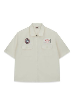 Chemise à manches courtes de couleur crème en coton, dotée de deux poches poitrine et de patchs brodés, avec une fermeture éclair et un col plat.