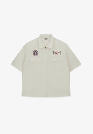 Chemise à manches courtes de couleur crème en coton, dotée de deux poches poitrine et de patchs brodés, avec une fermeture éclair et un col plat.