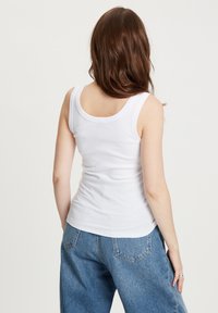 Biały, prążkowany top na ramiączkach z okrągłym dekoltem i szerokimi ramiączkami, noszony z luźnymi niebieskimi dżinsami. Widok z tyłu ukazuje gładką fakturę i prosty design.
