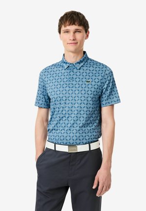 Giovane uomo che indossa una polo blu a maniche corte con motivo, pantaloni blu navy e cintura bianca, in piedi con una mano in tasca.