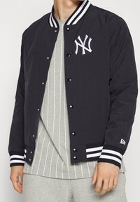 Svart bomberjacka med vita randiga ribbade manschetter och krage. Har en broderad NY-logotyp och tryckknappsstängning. Bärs över en grå randig skjorta.