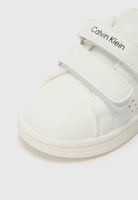 Baskets en cuir blanc avec deux sangles Velcro, bout rond, détails perforés sur le côté et semelle en caoutchouc. Logo affiché sur la sangle.