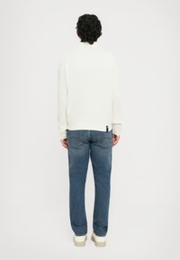 Muškarac s kovrčavom kosom, odjeven u bijeli pulover, plave traperice i bijele tenisice, stoji okrenut leđima prema jednostavnoj bijeloj pozadini.