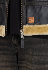 Jachetă de piele neagră pentru bărbați, cu margini de blană bej, fermoar frontal și patch cu logo; purtată peste un tricou negru și pantaloni din denim închis.