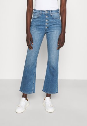 Personne portant un jean bleu taille haute avec des boutons à l'avant et des baskets blanches, debout devant un fond blanc uni.