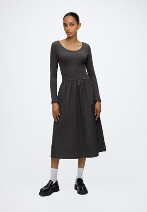 Frau trägt ein langärmeliges schwarzes Kleid mit geraffter Taille, weiße Socken und schwarze Plateau-Loafer und steht vor einem schlichten Hintergrund.