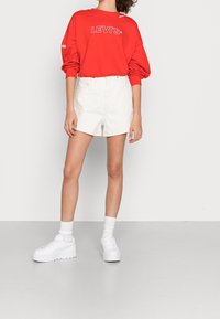 Felpa rossa con logo "LEVI'S", pantaloni corti bianchi a vita alta e sneakers bianche chunky con suola spessa. Calzini bianchi alla caviglia visibili.