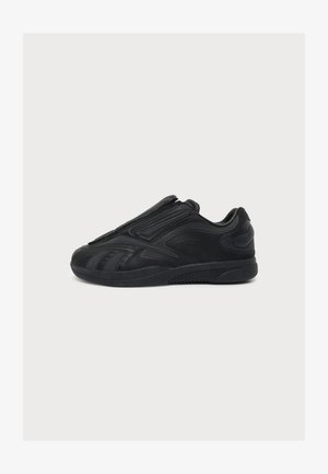 Zapatilla deportiva negra con diseño elegante, suela texturizada y estilo slip-on sin cordones visibles, mostrada de lado sobre un fondo blanco.