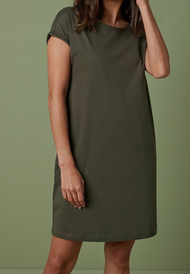 Femme portant une robe courte olive à manches courtes, longueur genoux, debout devant un fond vert assorti.