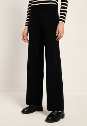 Lower body of person wearing black wide-leg pants, black loafers, and a black and white striped long-sleeve top against a neutral background.