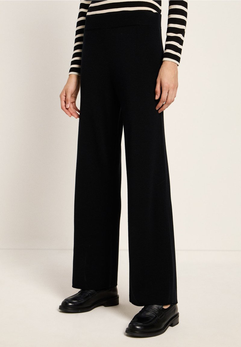 Unterkörper einer Person in schwarzen weiten Hosen, schwarzen Slippern und einem schwarz-weißen gestreiften Langarmoberteil vor einem neutralen Hintergrund.