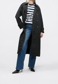 Abrigo largo negro tipo gabardina hecho de tela lisa, con botones cruzados delanteros. Lleva puesto sobre una camisa de rayas y combinado con jeans vaqueros de pierna ancha y tacones puntiagudos.