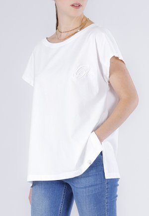 Kvinde iført løs hvid kortærmet top med diskret broderi på brystet og blå jeans, hånd i lommen, guldhalskæder synlige.